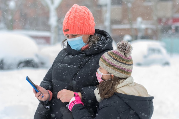 Madre e figlia indossano maschere di coronavirus che consultano il loro smartphone sotto un'intensa nevicata.