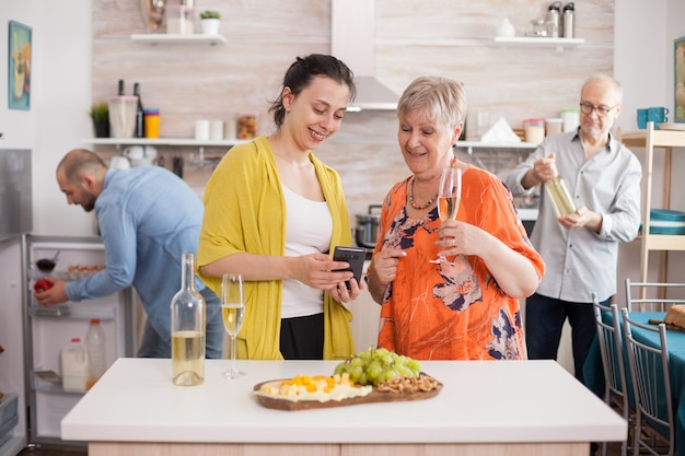 Madre e figlia navigano in internet utilizzando smarthphone in cucina durante la riunione di famiglia e bere vino.