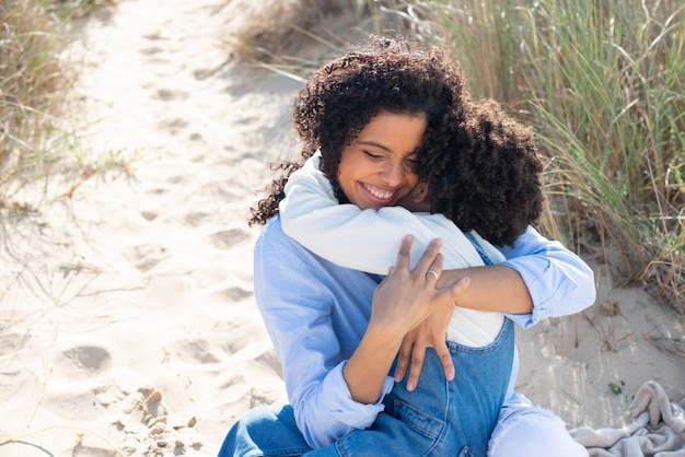 Madre e figlia premurose dell'afroamericano sulla spiaggia. Madre e bambino in abiti casual seduti su una coperta, abbracciati, con gli occhi chiusi. Famiglia, relax, concetto di natura