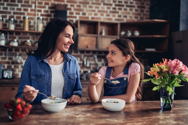 Madre e figlia sorridenti che si siedono al tavolo e mangiano fiocchi di mais colorati per colazione e si guardano in cucina