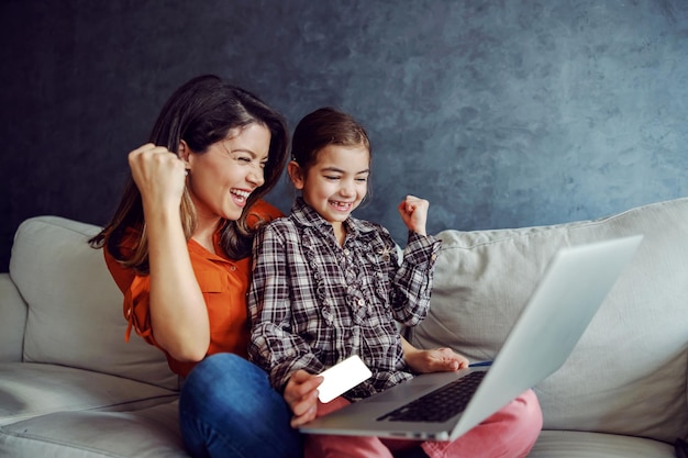 Madre e figlia sorridenti che si siedono sul divano e utilizzano il computer portatile per lo shopping online