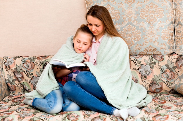 Madre e figlia stanno leggendo un libro sul divano di casa. una donna e un bambino sono seduti su un divano, avvolti in una morbida coperta e leggono un libro.