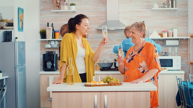 Madre e figlia tintinnano bicchieri di vino seduti in cucina. Famiglia allargata che festeggia nella sala da pranzo bevendo un bicchiere di vino mentre gli uomini cucinano in sottofondo