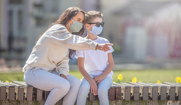 Madre e figlio in maschera siedono su una panchina della città con lei che indica la direzione in cui entrambi stanno guardando.