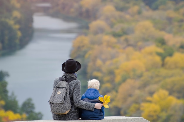 Madre e figlio sono seduti su un'altura. Autunno. Fiume e foresta sottostante. Vista posteriore