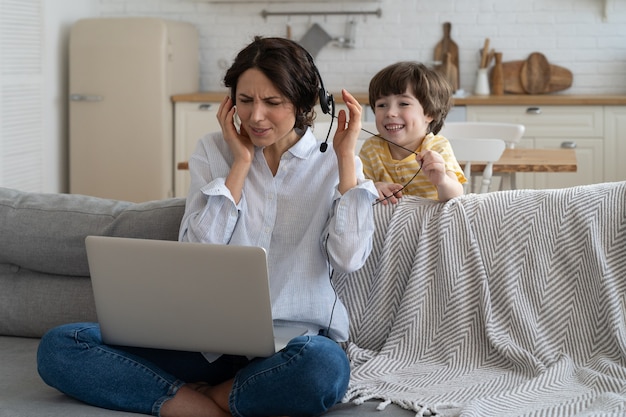 Madre esausta seduta sul divano a casa, lavorando al laptop, bambino distratto e chiedendo attenzione