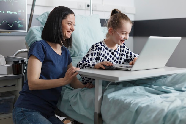 Madre felice allegra che si gode del tempo con la figlia sorridente mentre si trova nella stanza del reparto di clinica. Madre e figlia rilassate sedute nella stanza del reparto pazienti della clinica pediatrica mentre utilizzano un computer moderno.