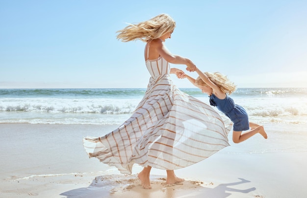 Madre felice che oscilla e fa girare la figlia carina in cerchio per le braccia in spiaggia Bambino giocoso, energico e gioioso che si diverte mentre si lega con la mamma durante le soleggiate vacanze estive all'aperto