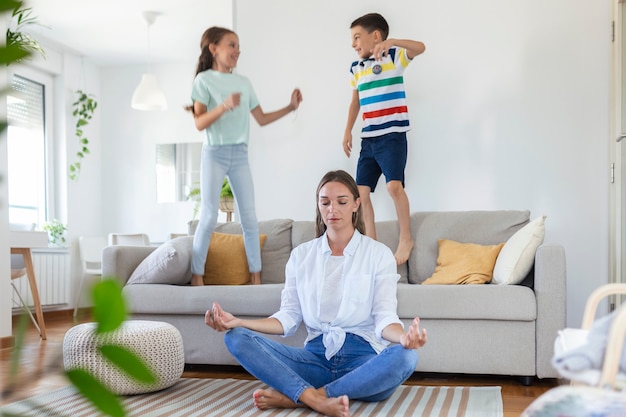 Madre felice con gli occhi chiusi che medita nella posa del loto sul pavimento cercando di salvare l'armonia interiore mentre i bambini eccitati saltano sul divano e urlano in un luminoso e spazioso soggiorno