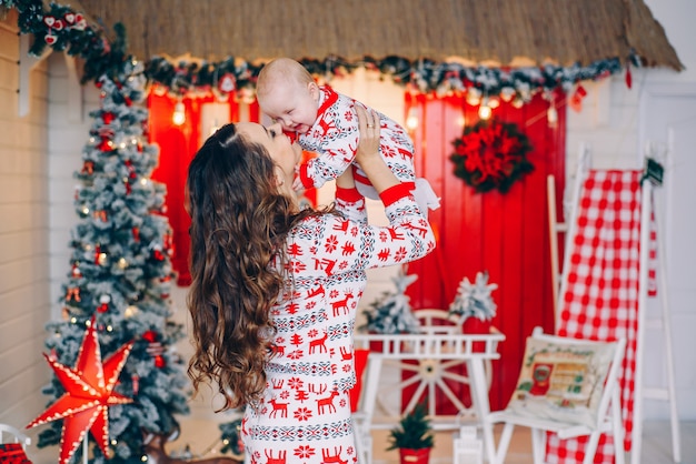 Madre felice con la sua piccola figlia in vestiti di festa con cervi e fiocchi di neve stampati nella stanza decorata con un albero di Natale e una corona