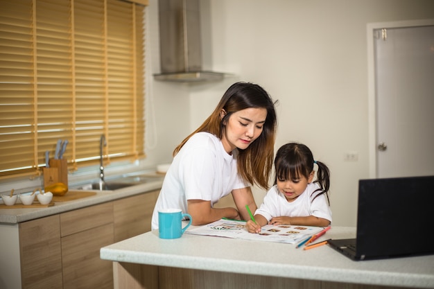 Madre felice e figlia sorridente che dipingono insieme usando i pennarelli. Madre che aiuta il bambino adottato con i compiti d'arte. Madre allegra e bambina asiatica che fanno pittura a casa.