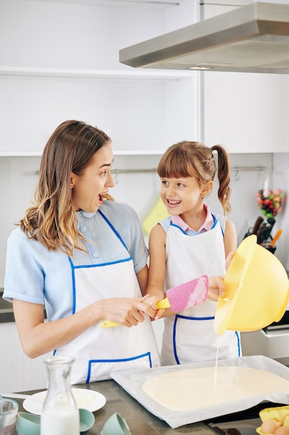 Madre felice eccitata e figlia preadolescenziale che si guardano l'un l'altro quando versano l'impasto liquido che hanno fatto sulla teglia