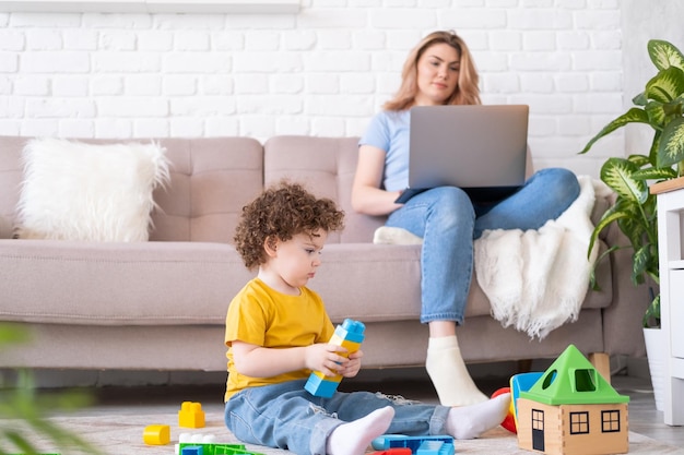 Madre freelance che utilizza laptop e telefono seduto sul divano con la figlia del bambino che gioca sullo smartphone