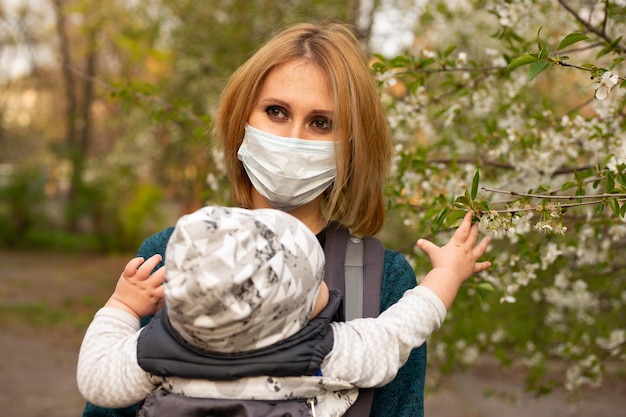 Madre in maschera medica con figlio piccolo nel parco cammina vicino a un albero bianco in fiore