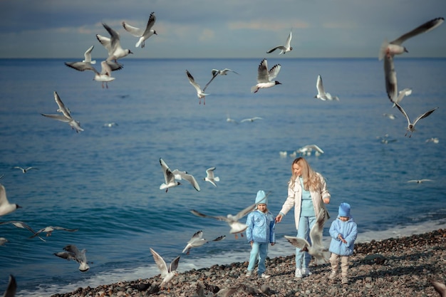 Madre in vacanza in famiglia con due figlie in abiti caldi durante una passeggiata in riva al mare che alimenta i gabbiani