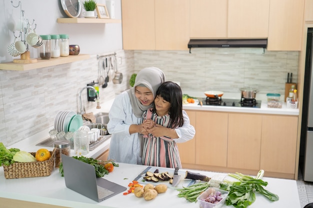 Madre musulmana che guarda la ricetta dal computer portatile e cucina con sua figlia. divertirsi donna con hijab e bambino che prepara la cena insieme