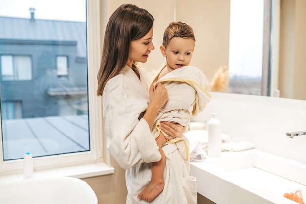 Madre soddisfatta in piedi vicino alla finestra del bagno ad ammirare il suo bambino carino in un accappatoio bianco