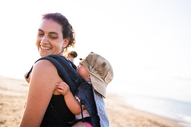 Madre sorridente che porta un bambino con un berretto sulla schiena in un marsupio all'aperto