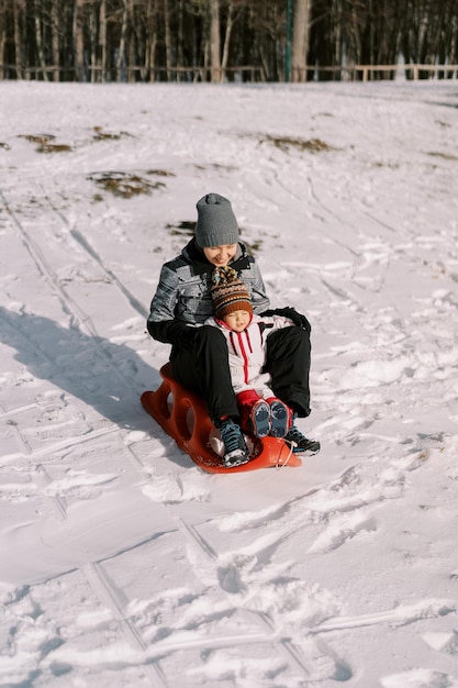 Madre sorridente con un bambino in slitta che scende da una collina coperta di neve