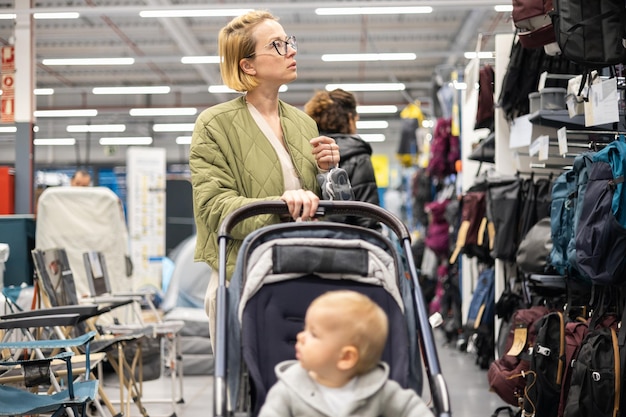 Madre vestita casualmente che sceglie scarpe sportive e prodotti di abbigliamento nel reparto sportivo del supermercato con il suo bambino neonato nel passeggino