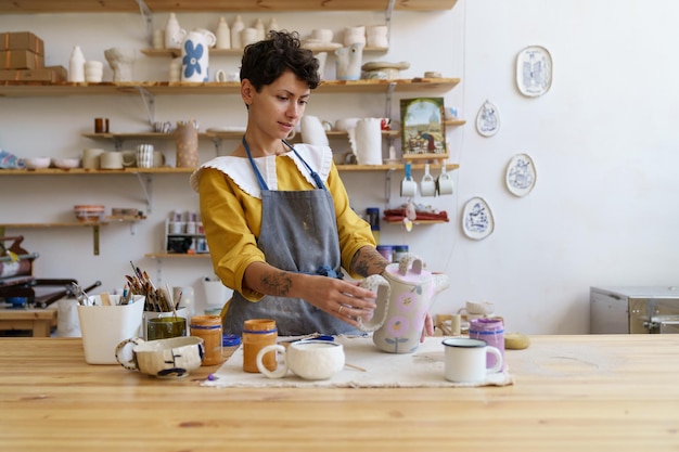 Maestra artistica della ragazza ceramista al lavoro donna felice in grembiule che tiene brocca fatta a mano in studio di ceramica