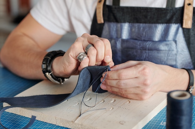 Maestro di artigianato creativo con ago che tiene piccolo pezzo di pelle scamosciata nera su tavola di legno sul tavolo mentre si cuce qualcosa