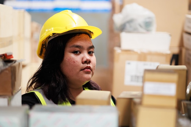 Magazziniere obeso asiatico in giubbotto di sicurezza e elmetto protettivo in fabbrica di magazzino prodotto industriale in scatole di cartone sugli scaffali Plus size femmina che ispeziona i prodotti durante il lavoro