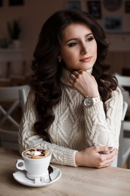 Magica incredibile bellezza di una giovane donna seduta in un caffè con una tazza di caffè e guardando fuori dalla finestra. Bruna sogna. Bei capelli mossi, trucco di lusso. Decorato con caffè art.