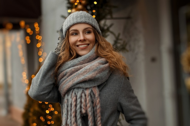 Magico ritratto felice di una giovane e bella donna sorridente in abiti a maglia vintage con un cappello e una sciarpa in vacanza vicino a luci gialle