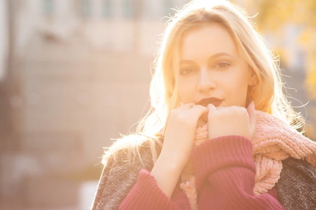 Magnifica donna bionda con labbra rosse che indossa sciarpa e cappotto lavorati a maglia, in posa sotto i raggi del sole. Spazio per il testo