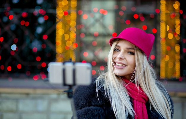 Magnifica ragazza bionda turistica che indossa un cappello divertente, che si fa selfie sulla strada della città decorata con ghirlande. Spazio per il testo