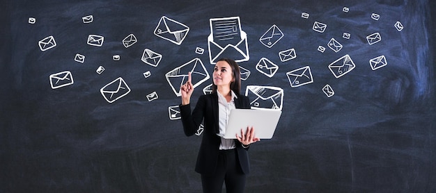 Mailing list e concetto di marketing con giovane donna con laptop su sfondo lavagna con buste e lettere