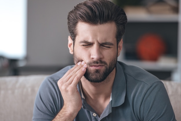 Mal di denti. Giovane frustrato che si tocca la guancia e tiene gli occhi chiusi mentre è seduto sul divano di casa