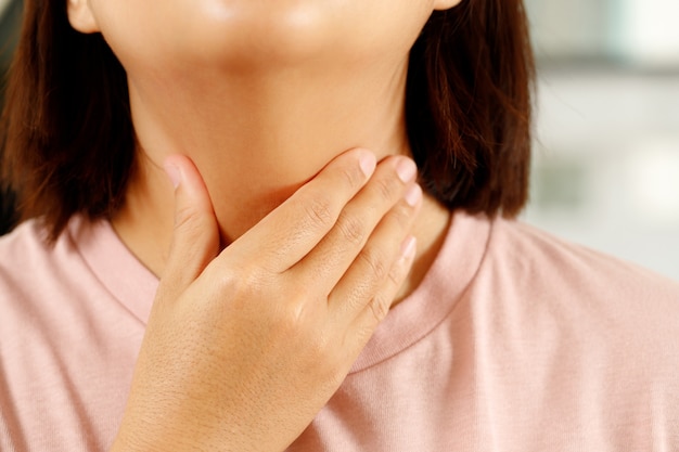 Mal di gola. Primo piano della giovane donna malata che tiene la sua gola infiammata usando le mani per toccare il collo malato in camicia blu su sfondo grigio. Concetto medico e sanitario. Concentrati sul rosso per mostrare il dolore.