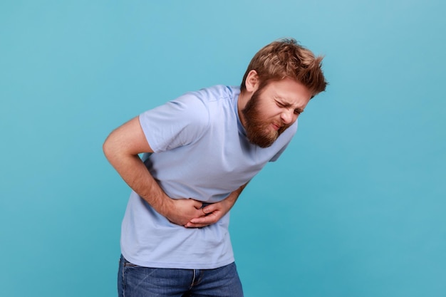 Mal di stomaco. Vista laterale dell'uomo barbuto in piedi e tenendo il ventre doloroso, sentirsi male, soffrire di diarrea o costipazione, rischio di appendicite. Studio indoor girato isolato su sfondo blu.