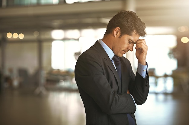 Mal di testa stress uomo d'affari in piedi e nel suo ufficio triste o sconvolto dal fallimento del debito sul lavoro Salute mentale o ansia burnout e lavoratore aziendale maschio esausto o depresso frustrato e stanco