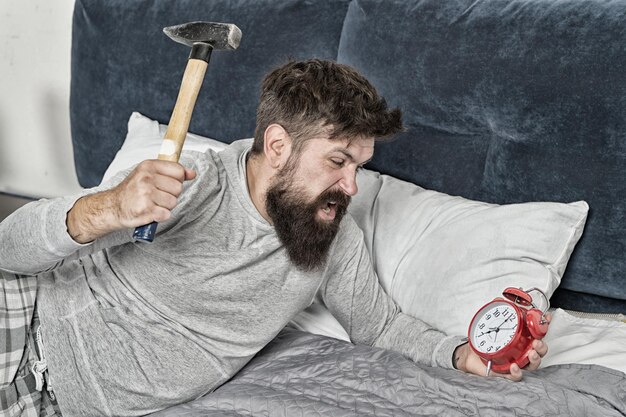 Malato e stanco Il ragazzo irritato distrugge l'orologio fastidioso Uomo sveglio insoddisfatto della sveglia che suona Dormi più a lungo Concetto di sonno sano uomo barbuto hipster vuole dormire odia il rumore della sveglia