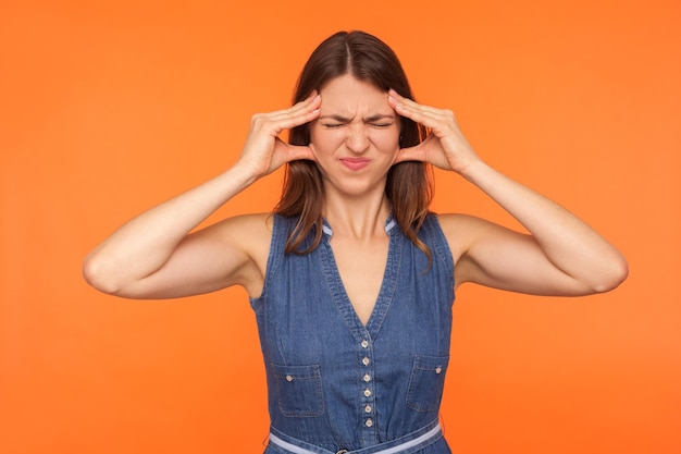 Male alla testa. Donna bruna malata depressa in abito di jeans che stringe la testa e fa una smorfia per un dolore terribile, soffre di emicrania, sintomi influenzali della febbre. tiro in studio indoor isolato su sfondo arancione