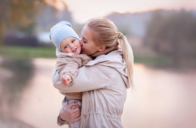 Mamma abbracci e baci sorridenti Bambino al tramonto sul lago natura. Sfondo sfocato, copia spazio, colore beige. La mamma ama suo figlio