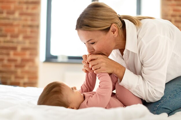 Mamma amorevole che tiene le mani della sua bambina e bacia svegliando la sua ghirlanda infantile dopo il sonno diurno sdraiato a letto