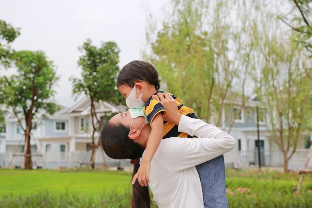 Mamma asiatica che porta suo figlio indossando una maschera protettiva e baciando nel giardino pubblico durante l'epidemia di coronavirus e influenza