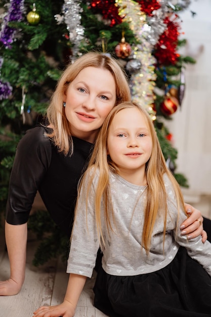 Mamma bionda e figlia che si siedono sotto l'albero di Natale. una felice madre single e una tradizione per festeggiare il nuovo anno a casa. l'atteggiamento di bambini e genitori.