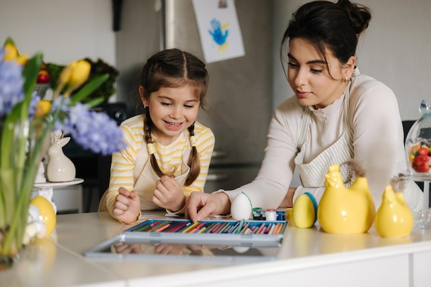 Mamma che aiuta sua figlia a dipingere una maschera da coniglietto pasquale Adorabile bambina seduta al tavolo in cucina