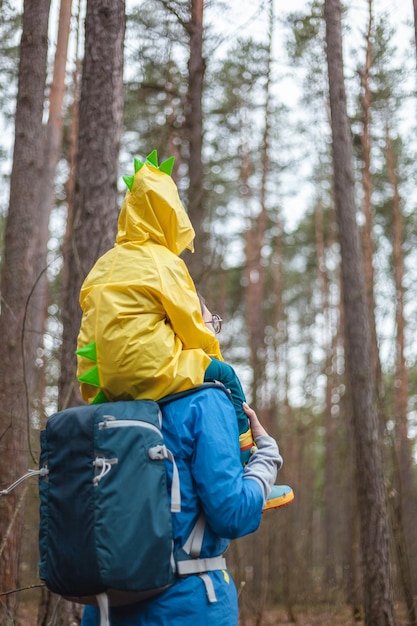 Mamma e bambino che camminano nella foresta dopo la pioggia in impermeabili insieme il bambino è seduto sulle spalle della vista posteriore della mamma