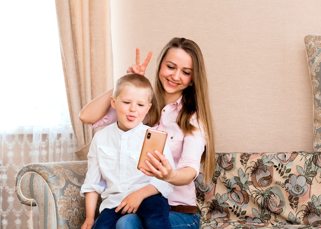 Mamma e bambino si fanno un selfie in un accogliente soggiorno. mamma e figlio fanno facce buffe e si fotografano davanti alla telecamera