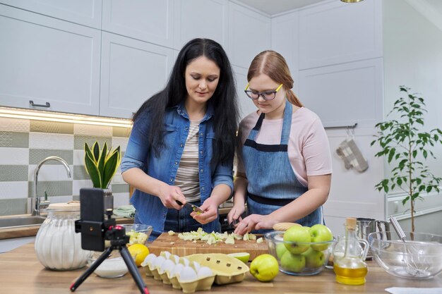 Mamma e figlia adolescente che cucinano insieme la torta di mele guardando lo schermo dello smartphone