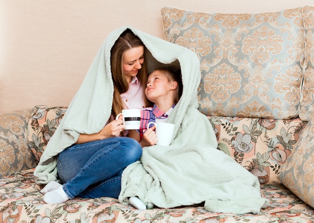 Mamma e figlia, avvolte in una morbida coperta, si siedono sul divano e bevono il tè.