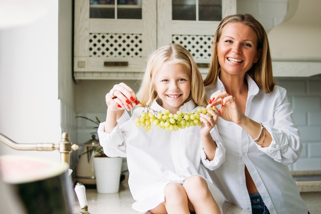 Mamma e figlia bionde felici dei capelli lunghi divertendosi con l'uva in cucina, stile di vita sano della famiglia