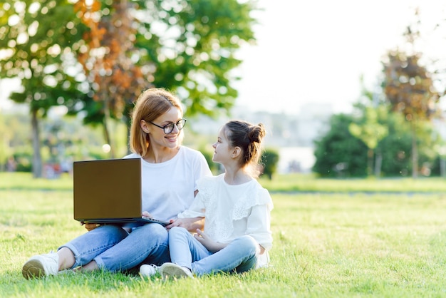 Mamma e figlia che giocano nel laptop all'aperto, ridendo e godersi il sole estivo sull'erba verde nel parco, famiglia all'aperto nel parco