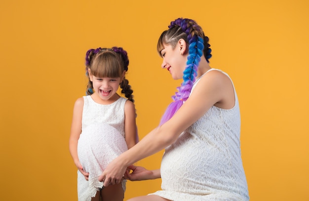 Mamma e figlia che ride positive con le stesse acconciature e abiti seduti sul pavimento in studio e in posa con una pancia incinta